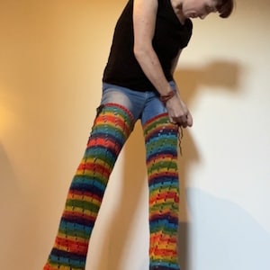 May include: A person wearing a pair of rainbow crochet leg warmers over blue jeans. The leg warmers are made with a colorful yarn and have a lacy pattern.