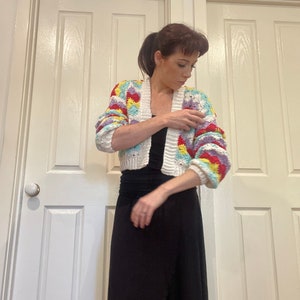 May include: A white crochet cardigan with colorful geometric patterns. The cardigan has a cropped length and long sleeves. The model is wearing a black maxi skirt.