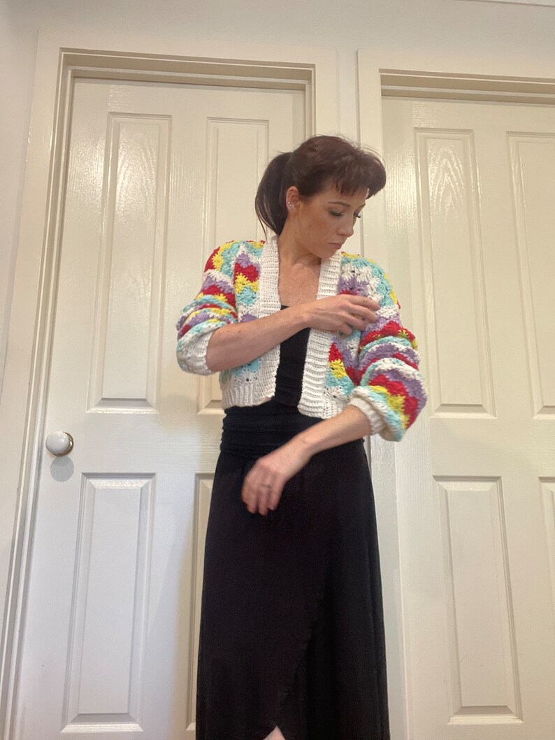 May include: A white crochet cardigan with colorful geometric patterns. The cardigan has a cropped length and long sleeves. The model is wearing a black maxi skirt.