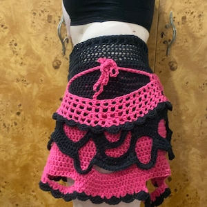 May include: A pink and black crocheted skirt with a layered design. The skirt has a scalloped hem and a tie at the waist.