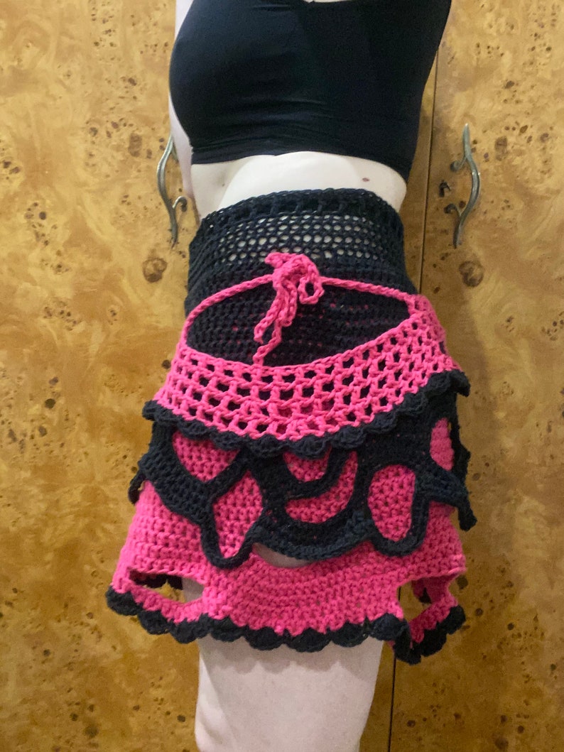 May include: A pink and black crocheted skirt with a layered design. The skirt has a scalloped hem and a tie at the waist.