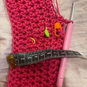 May include: A close-up of a pink crocheted fabric with a crochet hook and a measuring tape. The fabric has several colorful stitch markers.