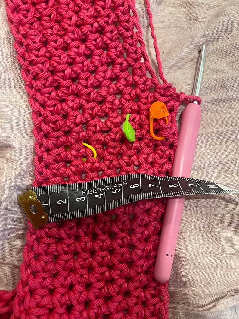 May include: A close-up of a pink crocheted fabric with a crochet hook and a measuring tape. The fabric has several colorful stitch markers.