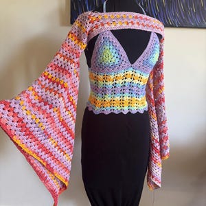 Op de afbeelding: Een kleurrijk gehaakt topje met lange, wijde mouwen. Het topje is gemaakt van een veelkleurig garen met roze, oranje, gele en paarse strepen. Het topje wordt gedragen over een zwarte rok.