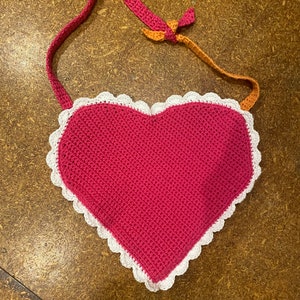 May include: A pink crocheted heart-shaped bag with a white crocheted trim and orange and brown straps.