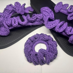 May include: A set of purple crochet sandals and a matching scrunchie. The sandals have black soles and a ruffled crochet design across the top. The scrunchie is also purple and has a ruffled design. The set is perfect for a casual, beachy look.