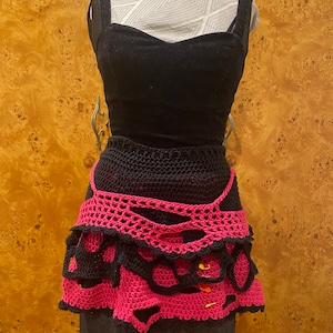 May include: A black and pink crocheted skirt with a layered design. The skirt has a scalloped edge and is decorated with small yellow safety pins.