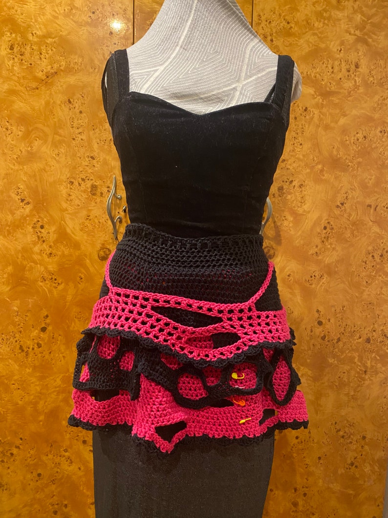 May include: A black and pink crocheted skirt with a layered design. The skirt has a scalloped edge and is decorated with small yellow safety pins.