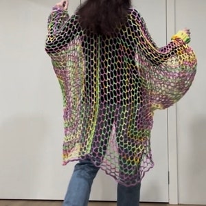 May include: A long, loose-fitting crochet cardigan in a rainbow color pattern. The cardigan is made of a lightweight, open-weave fabric and has a relaxed fit. It is shown being worn over blue jeans.