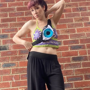 Può includere: Una donna indossa un top all'uncinetto con un grande disegno di occhio blu. Il top è verde, viola e bianco. Indossa pantaloni neri.