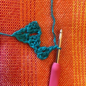 May include: Close-up of a teal crochet stitch being worked with a pink crochet hook. The crochet is being worked on a textured orange surface.