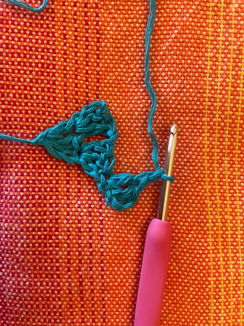 May include: Close-up of a teal crochet stitch being worked with a pink crochet hook. The crochet is being worked on a textured orange surface.