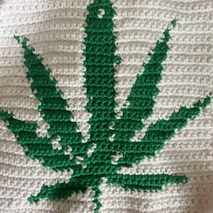 May include: A handmade crochet item featuring a large green cannabis leaf design on a white background. The leaf is centrally positioned, with detailed stitching defining its shape. The piece is likely a decorative item or part of a larger textile project.