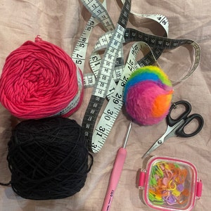 May include: A close-up of a craft project with pink and black yarn, a rainbow pom-pom, a pink crochet hook, a container of colorful stitch markers, and a pair of scissors.