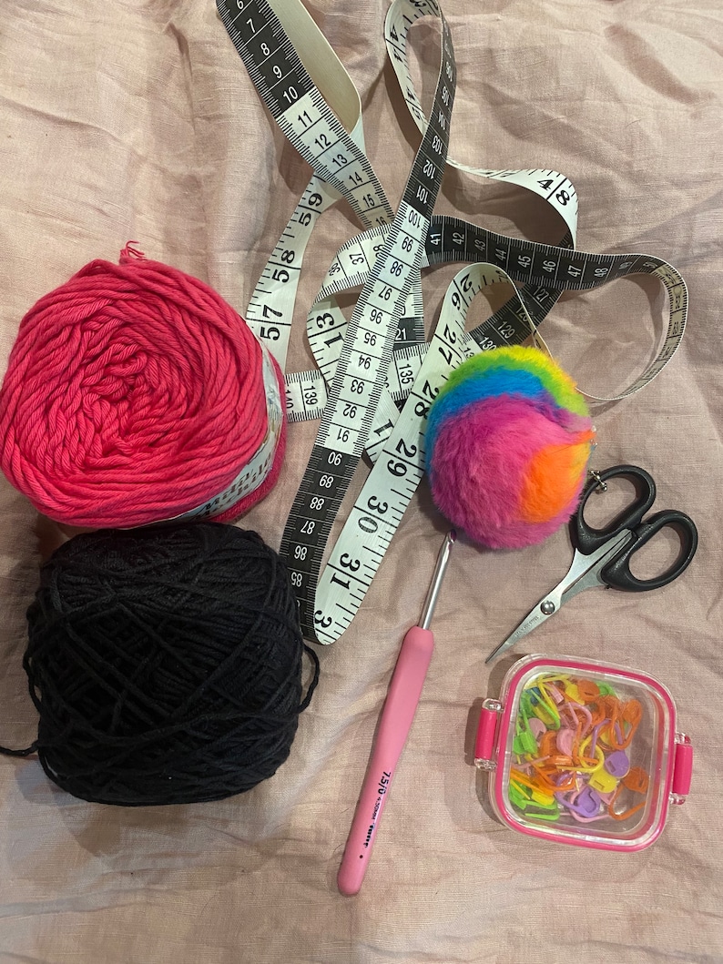 May include: A close-up of a craft project with pink and black yarn, a rainbow pom-pom, a pink crochet hook, a container of colorful stitch markers, and a pair of scissors.
