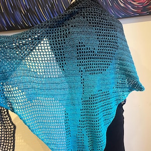 Op de afbeelding: Een teal en zwarte gehaakte sjaal met een kantpatroon. De sjaal is over een mannequin gehangen.