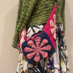 May include: A handmade crochet bag in pink, navy blue, and light pink. The bag features a large flower design on the front and a long black strap. The bag is worn over the shoulder.