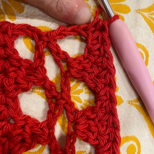May include: Close-up of red crochet fabric with a repeating pattern of circles. A pink crochet hook is visible in the upper right corner of the image.