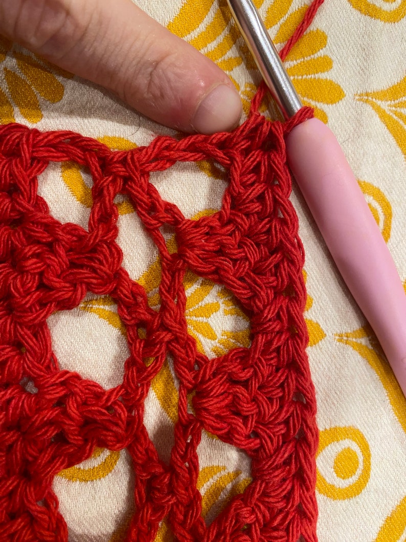 May include: Close-up of red crochet fabric with a repeating pattern of circles. A pink crochet hook is visible in the upper right corner of the image.
