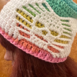 May include: A white crocheted hat with a rainbow stripe pattern. The hat is made with different colored yarns in shades of pink, yellow, green, and blue.