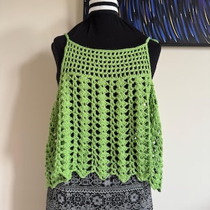 May include: A lime green crochet top with a square neckline and delicate straps. The open weave design creates a breezy, lightweight feel. The top is displayed on a black mannequin, paired with a patterned skirt. A piece of art hangs in the background.