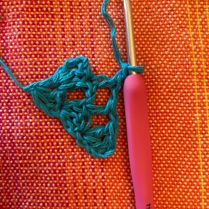 May include: A close-up of a teal crochet project in progress. A pink crochet hook with a metal hook is being used to work the stitches. The crochet hook is labeled "5.0 mm - J/10".
