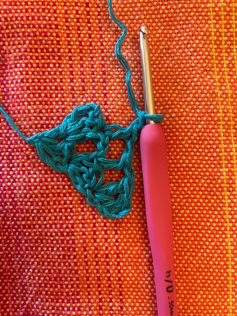 May include: A close-up of a teal crochet project in progress. A pink crochet hook with a metal hook is being used to work the stitches. The crochet hook is labeled "5.0 mm - J/10".