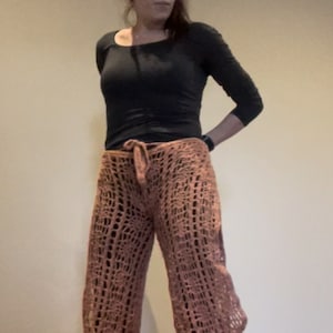 Puede incluir: Una mujer con una camisa negra de manga larga y pantalones de crochet marrones. Los pantalones tienen un corte holgado y fluido, con una pierna ancha.