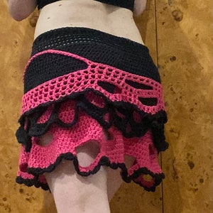 May include: A pink and black crocheted skirt with a layered design and cutouts. The skirt has a ruffled hem and is made with a delicate crochet stitch.