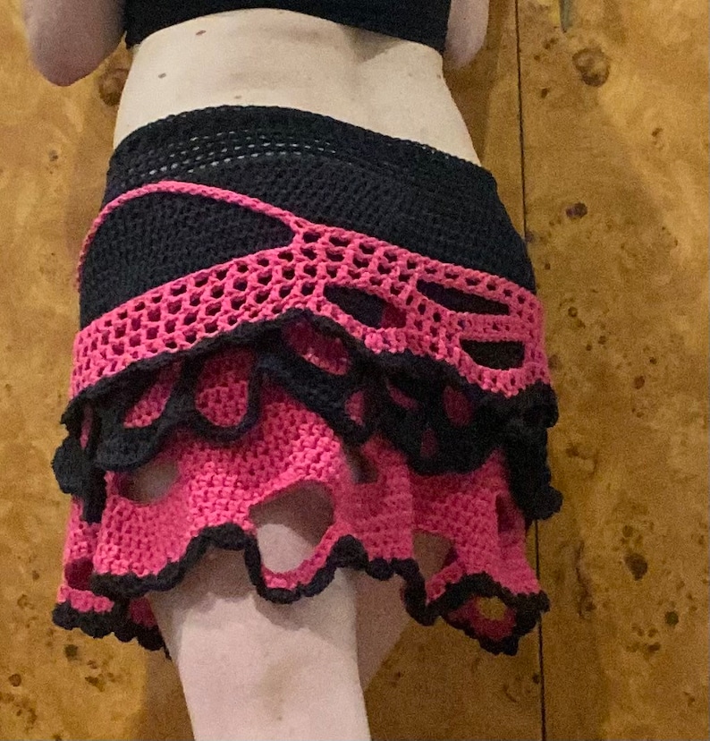 May include: A pink and black crocheted skirt with a layered design and cutouts. The skirt has a ruffled hem and is made with a delicate crochet stitch.