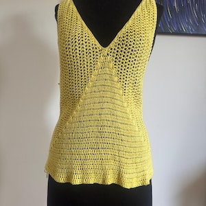 May include: A sleeveless, yellow crochet top with a deep V-neck. The open weave design creates a textured look. The top is displayed on a black mannequin, showcasing its shape and style. The garment's color is a bright, sunny yellow.
