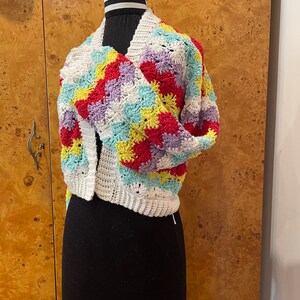 May include: A white crocheted cardigan with a colorful geometric pattern in shades of red, yellow, purple, and blue. The cardigan is shown on a mannequin.