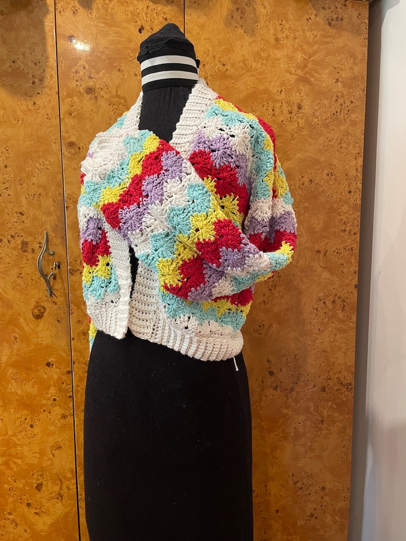 May include: A white crocheted cardigan with a colorful geometric pattern in shades of red, yellow, purple, and blue. The cardigan is shown on a mannequin.