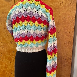 May include: A colorful crocheted sweater with a chevron pattern in shades of red, yellow, blue, and purple. The sweater has long sleeves and a cropped length.