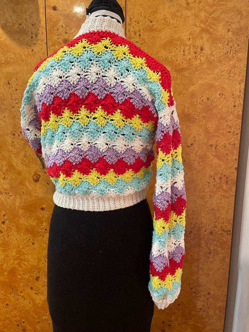 May include: A colorful crocheted sweater with a chevron pattern in shades of red, yellow, blue, and purple. The sweater has long sleeves and a cropped length.