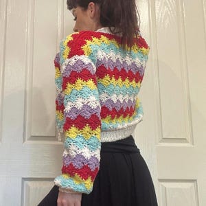 May include: A colorful crocheted sweater with a chevron pattern in shades of red, yellow, blue, white, and purple. The sweater is cropped and has long sleeves.