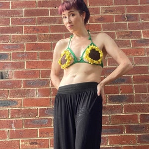 May include: A woman wearing a green and yellow crocheted sunflower bikini top. The top has a halter style and is made with yarn in shades of green and yellow. The sunflower design is crocheted in detail with brown centers and yellow petals.