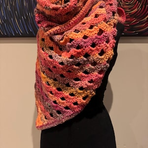 May include: A crocheted shawl in shades of orange, pink, and purple. The shawl is made with a lacy stitch pattern and has a triangular shape.