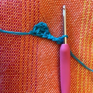 May include: A close-up of a turquoise crochet stitch being worked with a pink crochet hook. The hook is labeled "6/0".