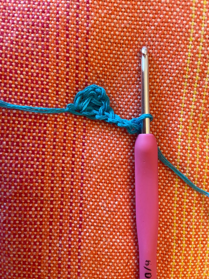 May include: A close-up of a turquoise crochet stitch being worked with a pink crochet hook. The hook is labeled "6/0".