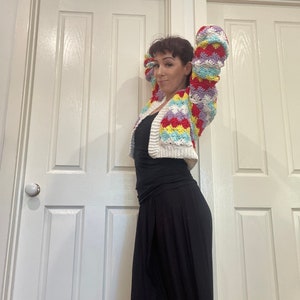 May include: A woman wearing a colorful crocheted cardigan with a white background and a black skirt.