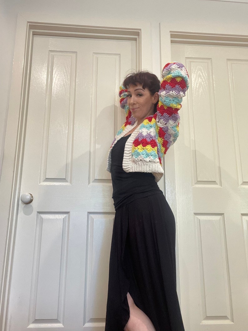 May include: A woman wearing a colorful crocheted cardigan with a white background and a black skirt.
