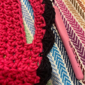May include: Close-up of a pink crochet hook with a black crochet border on a pink and black striped fabric.