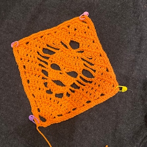 Puede incluir: Cuadrado de crochet naranja con un diseño de calavera. El cuadrado está sujeto a una superficie gris oscuro con imperdibles.