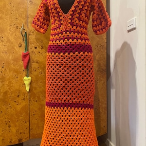 May include: A long, crocheted dress with a deep V-neckline. The dress is made in a multi-coloured pattern of orange, red, and pink.