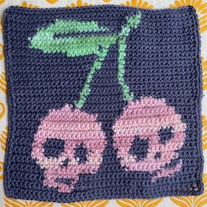 Op de afbeelding: Gehaakte vierkant met een donkerblauwe achtergrond, met een ontwerp van twee roze schedelkersen en groene bladeren. De schedels hebben een verloop van roze tinten. Het vierkant is gemaakt met een strakke haaksteek.