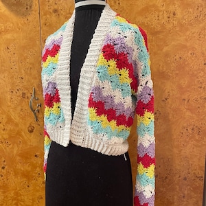 May include: A white crocheted cardigan with colorful stripes of red, yellow, blue, and purple. The cardigan has a V-neck and long sleeves.