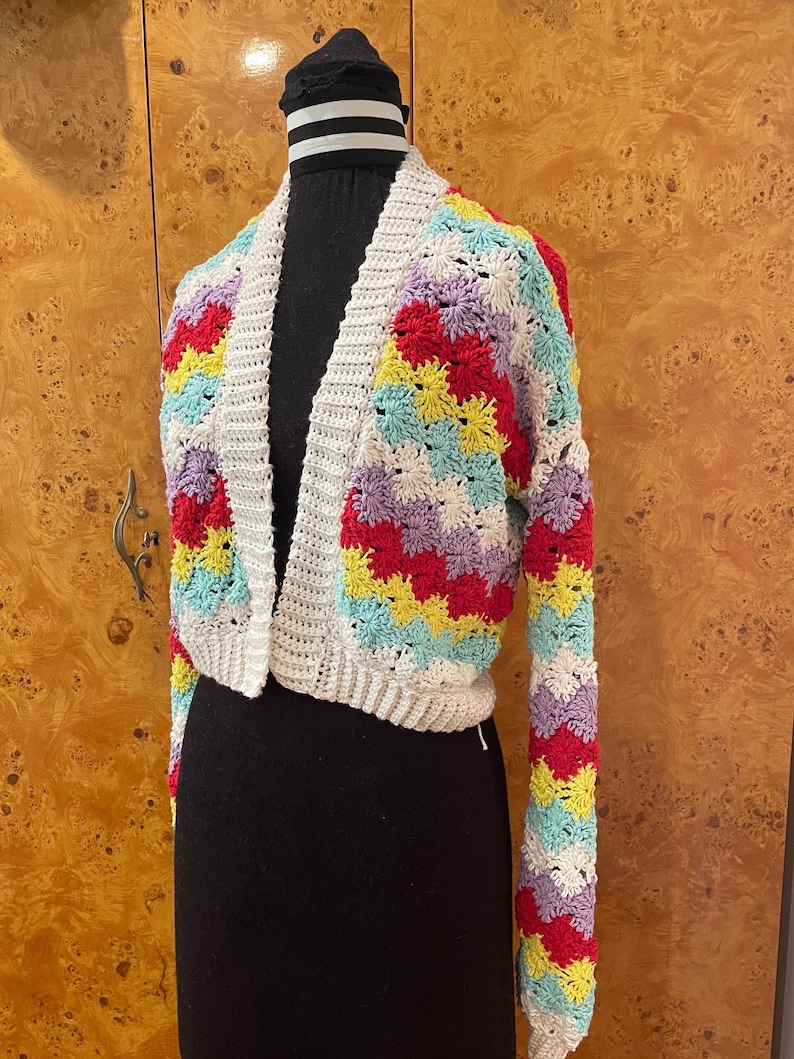 May include: A white crocheted cardigan with colorful stripes of red, yellow, blue, and purple. The cardigan has a V-neck and long sleeves.