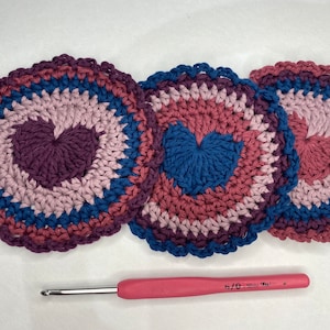 May include: Three crocheted coasters with a heart design in shades of pink, purple, and blue. The coasters are made with a textured stitch and have a scalloped edge. A pink crochet hook is also pictured.