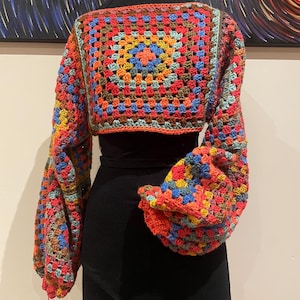 May include: A colourful crochet top with a square pattern and long, puffy sleeves. The top is a cropped style and is made with a variety of colours, including red, orange, yellow, blue, and green.